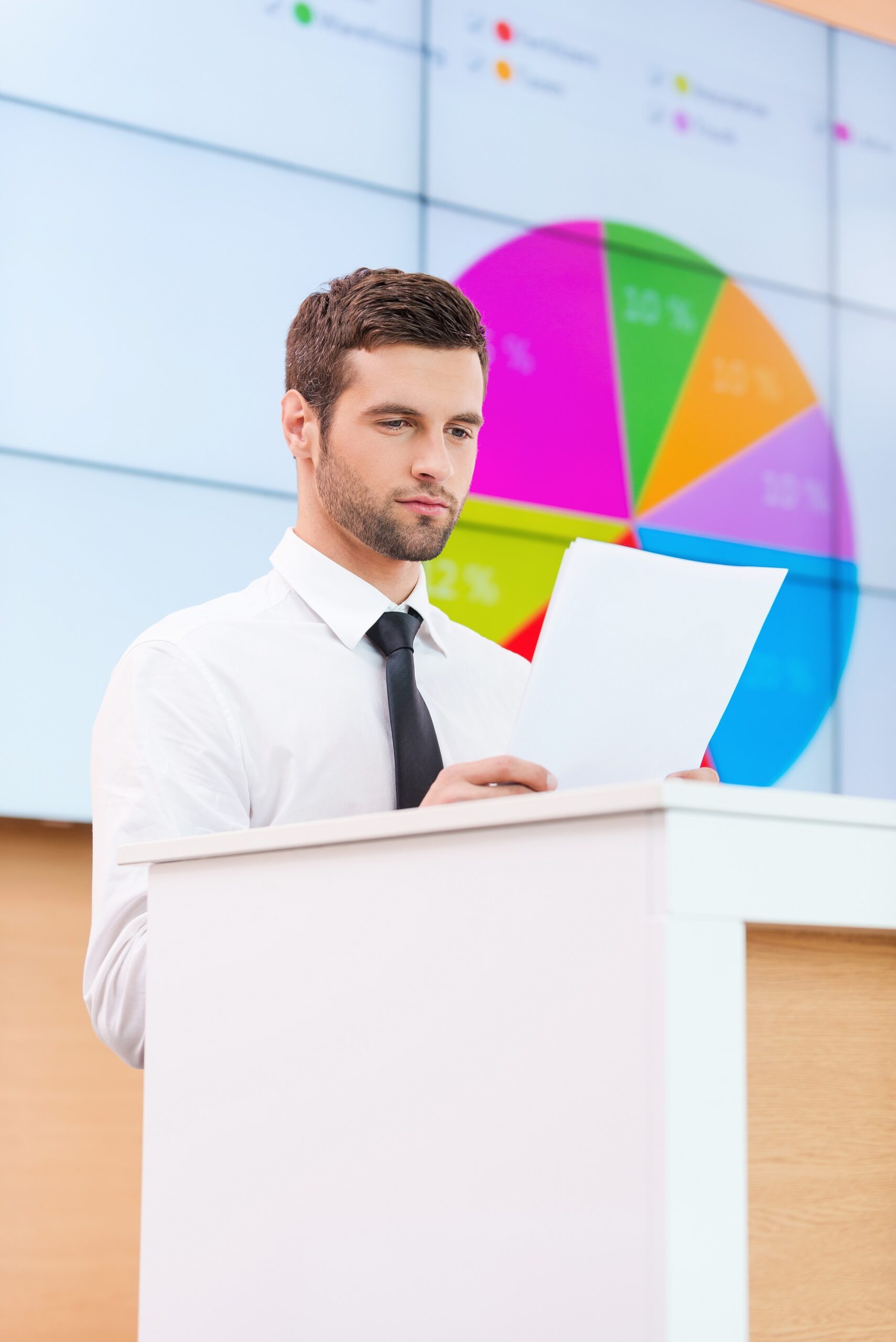 Getting ready to presentation. Confident young man in formalwear standing at the tribune and looking at paper while making a presentation with projection screen in the background